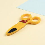 Yellow bird-design safety scissors lie closed on a pale cream surface beside a light blue sheet of paper.