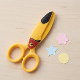 Yellow bird-design safety scissors rest on a wooden table beside pastel paper cutouts shaped as a flower, star, and circle.