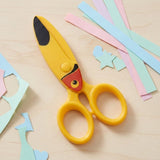 Yellow safety scissors with a red and black bird design rest on light wood beside pastel paper strips and cutouts.