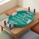 A green tabletop soccer game with grey goal ends and dark control handles sits on a wooden table surrounded by floor cushions.