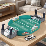 A tabletop soccer/football game set displayed on a light wood table in a living room setting. The product includes a green soccer field oval, gray and white goal structures with black handles, small black-and-white soccer balls, and a visible 'FOOTBALL' logo on the front.