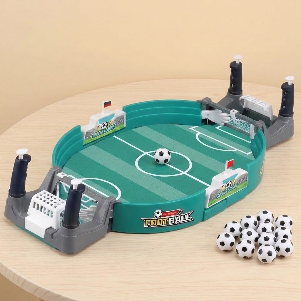 A children's tabletop soccer game set on a light wood table against a beige background. The oval green field toy has white markings, gray goals with black posts, small white flags, and several black-and-white soccer balls.
