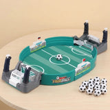 A children's tabletop soccer game set on a light wood table against a beige background. The oval green field toy has white markings, gray goals with black posts, small white flags, and several black-and-white soccer balls.