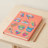 Diamond painting kit stickers decorate a coral notebook beside a wooden pencil on a pale wood table.