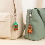 Two cute cartoon animal zipper keychains hanging from bags. On the left, a green alligator charm with a brown bear inside hangs from a beige canvas backpack.
