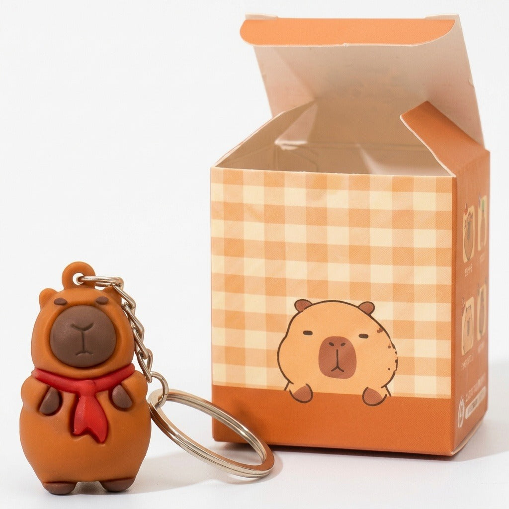 A cute capybara keychain charm in brown with a red scarf, shown next to its orange and beige checkered packaging box on a white background.