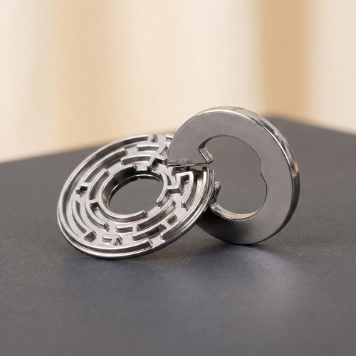 Silver metal brain teaser puzzle with interlocking circular maze pieces on a dark gray surface against a blurred beige background.
