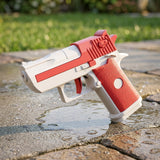 A red-and-white water gun with a textured red grip stands on wet stone paving beside a blurred green lawn.