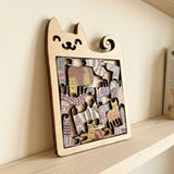 Wooden cat-shaped jigsaw puzzle with multicoloured patterned cat pieces leans against a cream wall on a light wooden shelf.