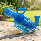 A transparent blue electric water gun with a top handle and lower reservoir rests on wet stone paving in a sunny garden.