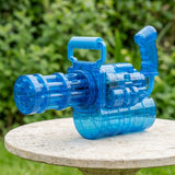 A transparent blue electric water gun with a top handle rests on a round stone table against green foliage.