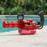 A transparent red electric water gun with black handles rests on pale paving beside a swimming pool.