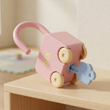 A pink car-shaped lock puzzle with cream wheels, an open shackle, and an inserted pale blue key rests on a sunlit wooden shelf.