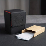 Black textured card deck box with red edging and embossed symbols beside an open cardboard sleeve containing clear card sleeves on dark fabric.
