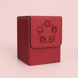 A red textured card deck box with a flap closure featuring a dark red circular emblem of four elemental symbols, set against a solid pale pink background.