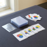 Clear card sleeves stacked beside loose and sleeved cards with colorful geometric designs on a dark tabletop near a window.