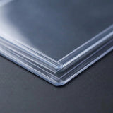 Clear PVC card sleeves are stacked on a dark gray textured surface, with their layered edges and corners shown close up.