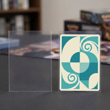 Clear PVC card sleeves stand on a dark tabletop, one empty and another holding a teal and cream patterned card.
