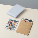 A stack of clear protective sleeves sits on a light beige surface next to a printed art card and a kraft paper sleeve holding another card. The art features a geometric face illustration in muted teal, beige, orange, and gray tones.