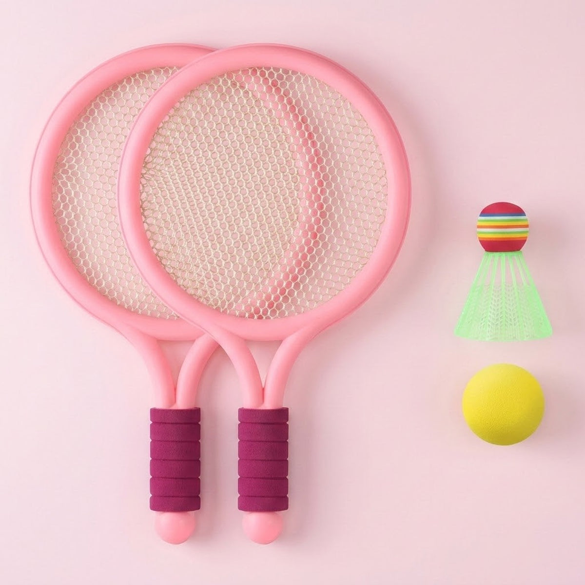 Pink plastic toy racket set with purple grips, white mesh, a yellow ball and green shuttlecock on a pale pink background.