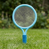 Blue plastic toy racket with white mesh and a ribbed blue grip stands upright in grass against blurred garden greenery.
