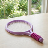 Purple plastic toy racket with white mesh and a magenta ribbed grip lies on a pale wooden table beside a garden-facing window.