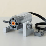 Gray building block accessory motor with an orange axle opening, black cable, and supported gear rests on a light gray surface.