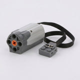 Grey plastic motor accessory for building block sets with an orange centre hub and a flat black cable, photographed on white.