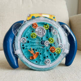 Round maze puzzle with blue handles, turquoise tracks and colorful gears beneath a transparent cover, resting on a cream upholstered seat.