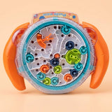 A children's toy puzzle maze with a circular clear plastic base, orange side handles, and colorful rotating gears in blue, green, orange, and dark blue. The toy is photographed on a plain warm beige background.