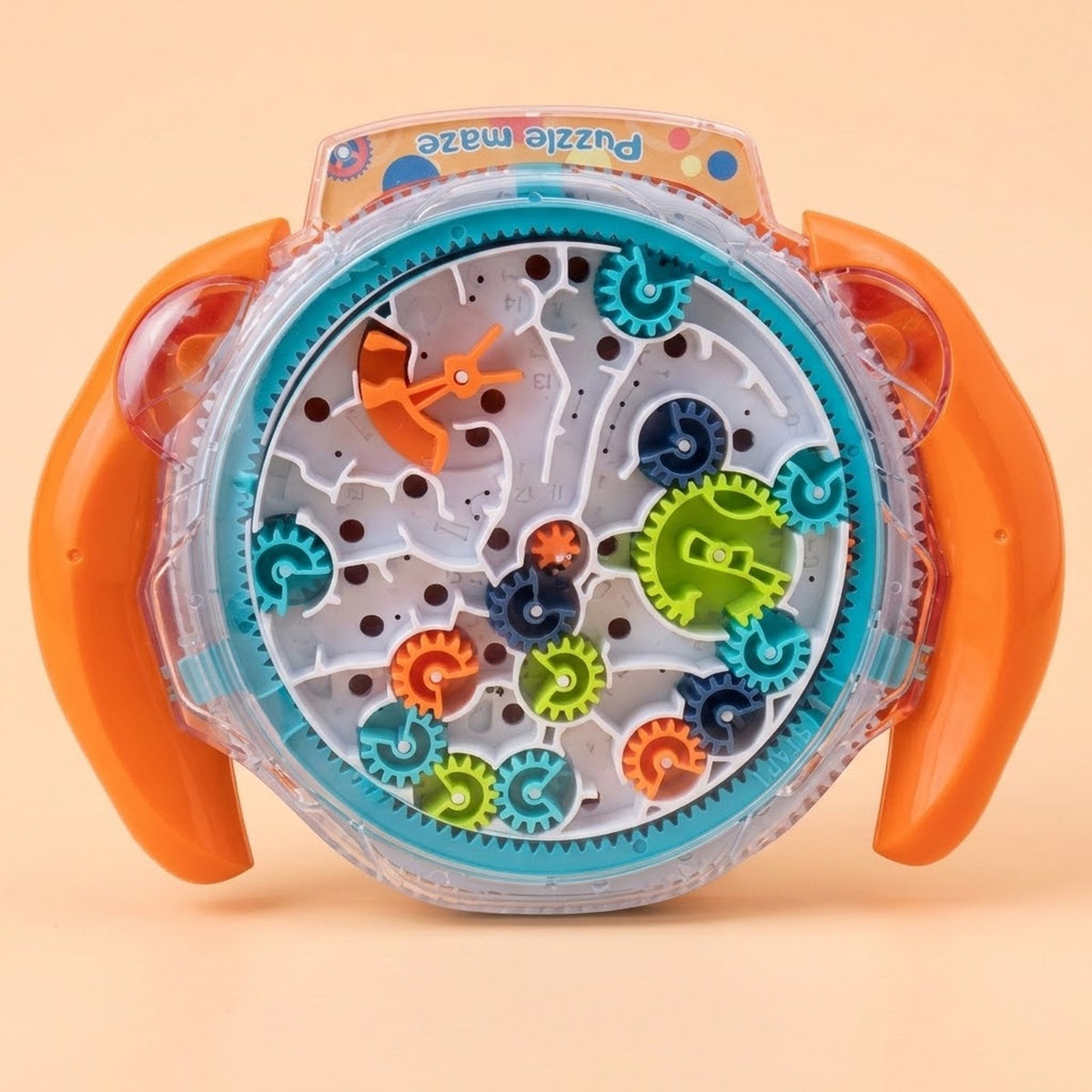 A children's toy puzzle maze with a circular clear plastic base, orange side handles, and colorful rotating gears in blue, green, orange, and dark blue. The toy is photographed on a plain warm beige background.