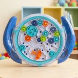 A circular maze puzzle with blue handles, a clear cover and colourful gears rests on a wooden table in front of colourful shelves.
