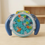 A maze puzzle with blue handles, turquoise tracks and colourful gears beneath a clear cover stands on a wooden table against a beige background.
