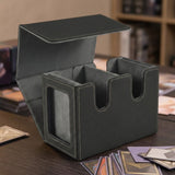 An open black-and-gray deck box with two empty compartments sits on a wooden table surrounded by scattered cards.