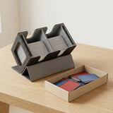 Black deck box with gray lining holds cards on a light wood table beside a cardboard tray of red and blue sleeves.