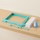 Penguin balance game pieces on a wooden shelf: turquoise plastic frame, white blocks, blue penguin figure, and two small shovel tools.