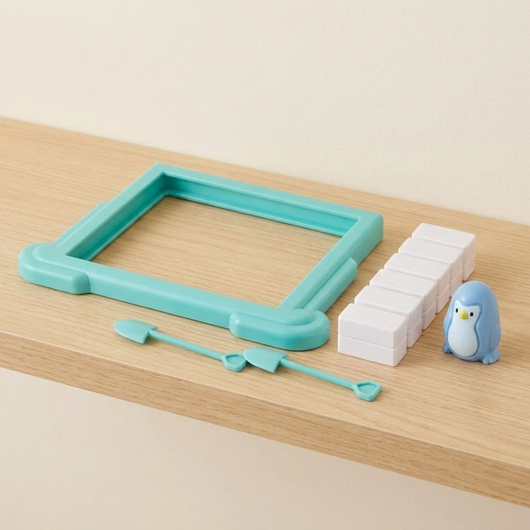 Penguin balance game pieces on a wooden shelf: turquoise plastic frame, white blocks, blue penguin figure, and two small shovel tools.