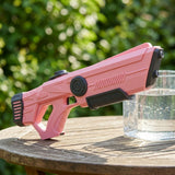Pink water gun with black accents rests on a wooden garden table beside a glass of water, with blurred greenery behind.