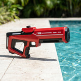 A red and black handheld water gun lies on the edge of a swimming pool, with water spraying from the nozzle. The background shows a light stone deck and blurred green foliage and pool water.