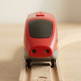 A red toy train with a dark windshield and gray wheels, positioned on a wooden track against a soft-focus light background.