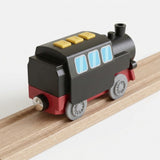 A small wooden toy train locomotive with a black body, red undercarriage, gray wheels, light blue windows, and yellow buttons, shown on a wooden rail against a clean white background.