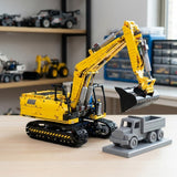 A yellow and black building-block excavator toy is displayed on a light wood table, lifting a gray toy dump truck with its bucket. The scene is set in a room with shelves of other toy vehicles in the background.