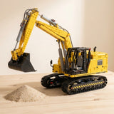Yellow and black plastic excavator building set assembled with its bucket raised beside a small sand pile on a wooden tabletop.