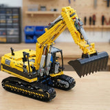 A yellow and black building block excavator toy with a black bucket, displayed on a light wooden tabletop with a blurred workshop background.