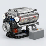 Silver plastic V8 engine model kit with transparent panels stands beside a gray control box against a light gray background.