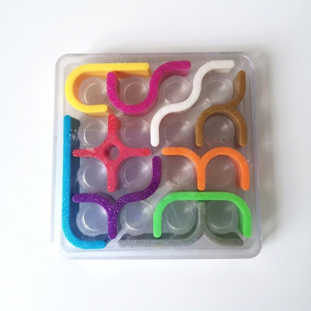 Mechanical puzzle viewed from above, with multicolored glittery plastic pieces arranged in a clear square tray against a white background.
