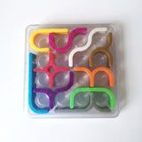 Mechanical puzzle viewed from above, with multicolored glittery plastic pieces arranged in a clear square tray against a white background.