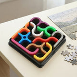 A mechanical puzzle with colorful curved plastic pieces in a black tray sits on a white tabletop beside scattered jigsaw pieces.