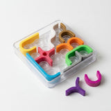 Mechanical puzzle in a clear tray on a white surface, with purple and pink curved pieces placed outside the tray.