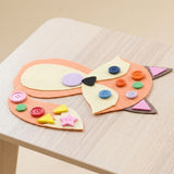 Craft kit project example showing a peach felt fox decorated with colorful buttons and foam shapes on a light wood tabletop.